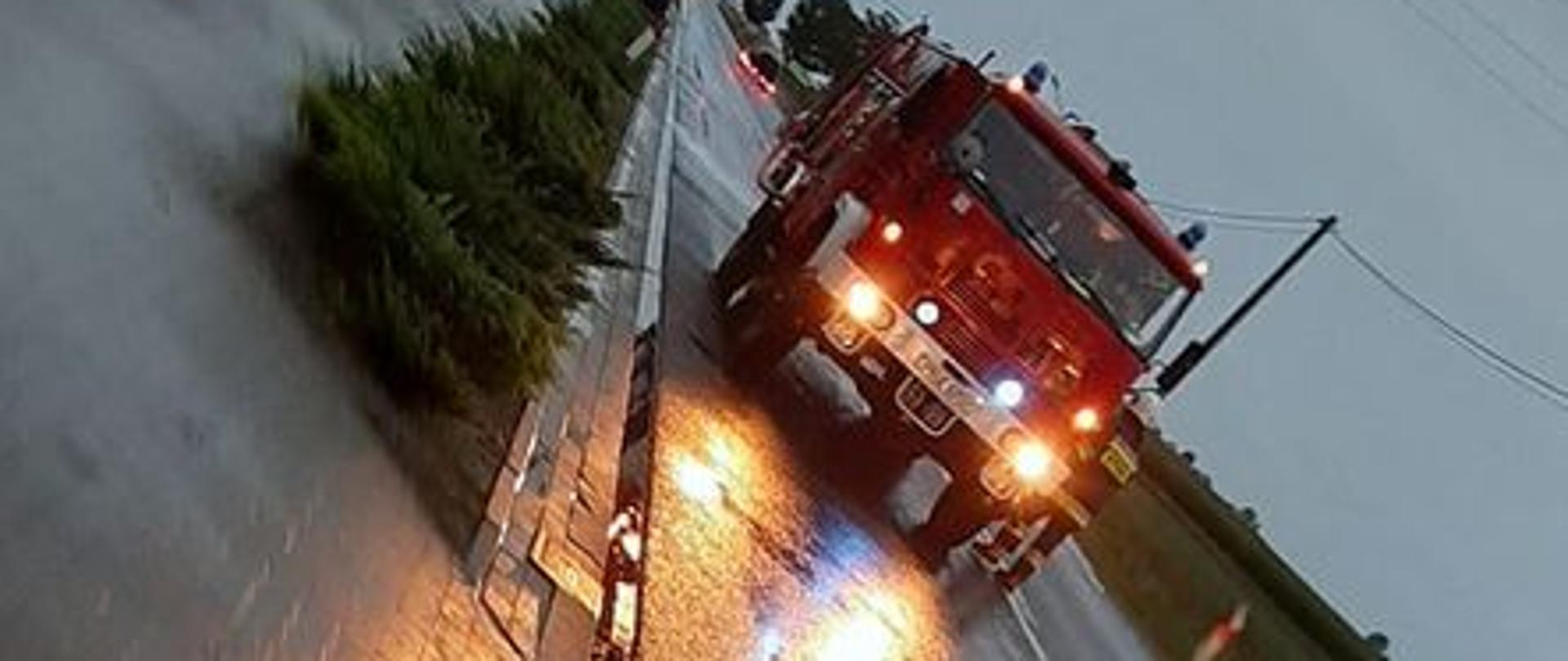 Na drodze stoi wóz strażacki z włączonymi światłami alarmowymi. Przed wozem na jezdni widoczny pas substancji ropopochodnej. W oddali widoczne inne samochody przemieszczające się w obu kierunkach. Z lewej strony chodnik oddzielony od jezdni iglakami oraz zabudowania. W poprzek drogi przebiega napowietrzna linia energetyczna.