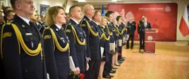Szereg funkcjonariuszy Państwowej Straży Pożarnej w galowych mundurach, trzymających w rękach róże i dokumenty, stoi na baczność w sali. W tle na podium przemawia mężczyzna.