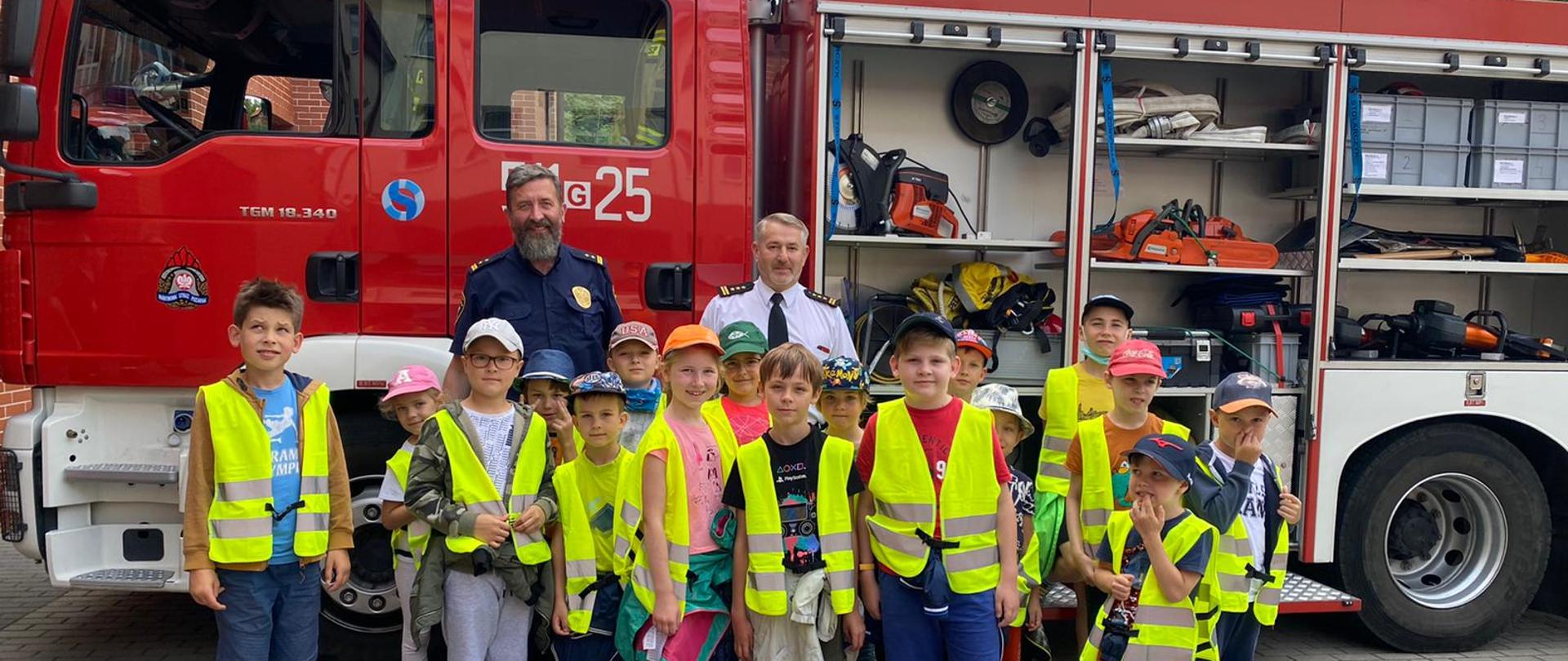 Dzieci ze szkoły karate z wizytą. Stoją w grupce w odblaskowych kamizelkach i pozują do zdjęcia. Za nimi samochód strażacki z otwartymi skrytkami oraz komendant straży pożarnej i straży miejskiej. Przed nimi na ziemi położona jest przecinarka tarczowa do stali i betonu.