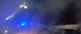 Na zdjęciu w porze nocnej widać palące się śmieci, głownie tworzywa sztuczne. Na zdjęciu panuje silne zadymienie. W zadymieniu widać strażaków gaszących pożar z kosza drabiny z wysokości kilkunastu metrów. W gęstym dymie na drugim planie widać samochód ratowniczo-gaśniczy.