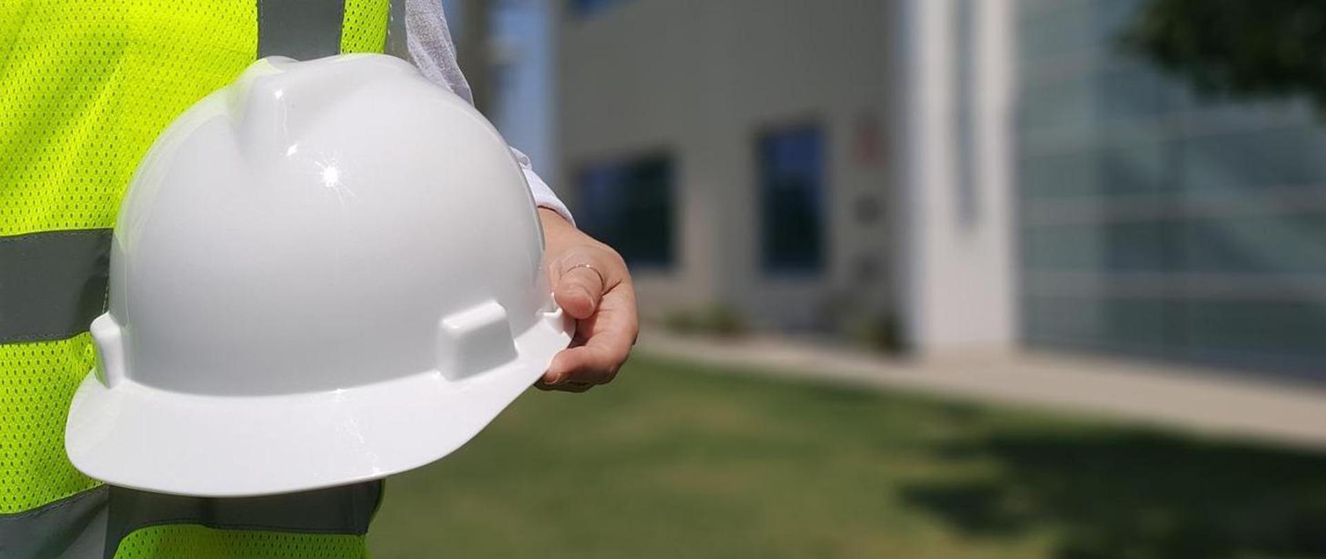 Osoba w kamizelce odblaskowej, trzymająca kask ochronny. Zbliżenie, widoczny tylko fragment postaci.