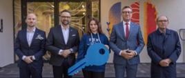 Przed wejściem do budynku stoi pięć osób, od lewej mężczyzna w granatowym garniturze, minister Kulasek, młoda dziewczyna w granatowiej bluzie z napisem UWM trzymająca wielki niebieski symboliczny klucz i dwóch mężczyzn w garniturach.