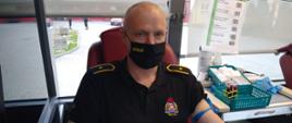 Zdjęcie przedstawia funkcjonariusza PSP, który oddaje krew w mobilnym autobusie krwiodawstwa. Strażak ma na twarzy maseczkę z napisem straż. Po jego prawej stronie widać różne przybory oraz przyrządy, które służą do pobierania krwi. W jego lewym przedramieniu znajduje się wkłucie przez które pobierana jest krew. W tle zdjęcia przez znajdującą się szybę widać stojących dwóch strażaków oraz samochód osobowy PSP.