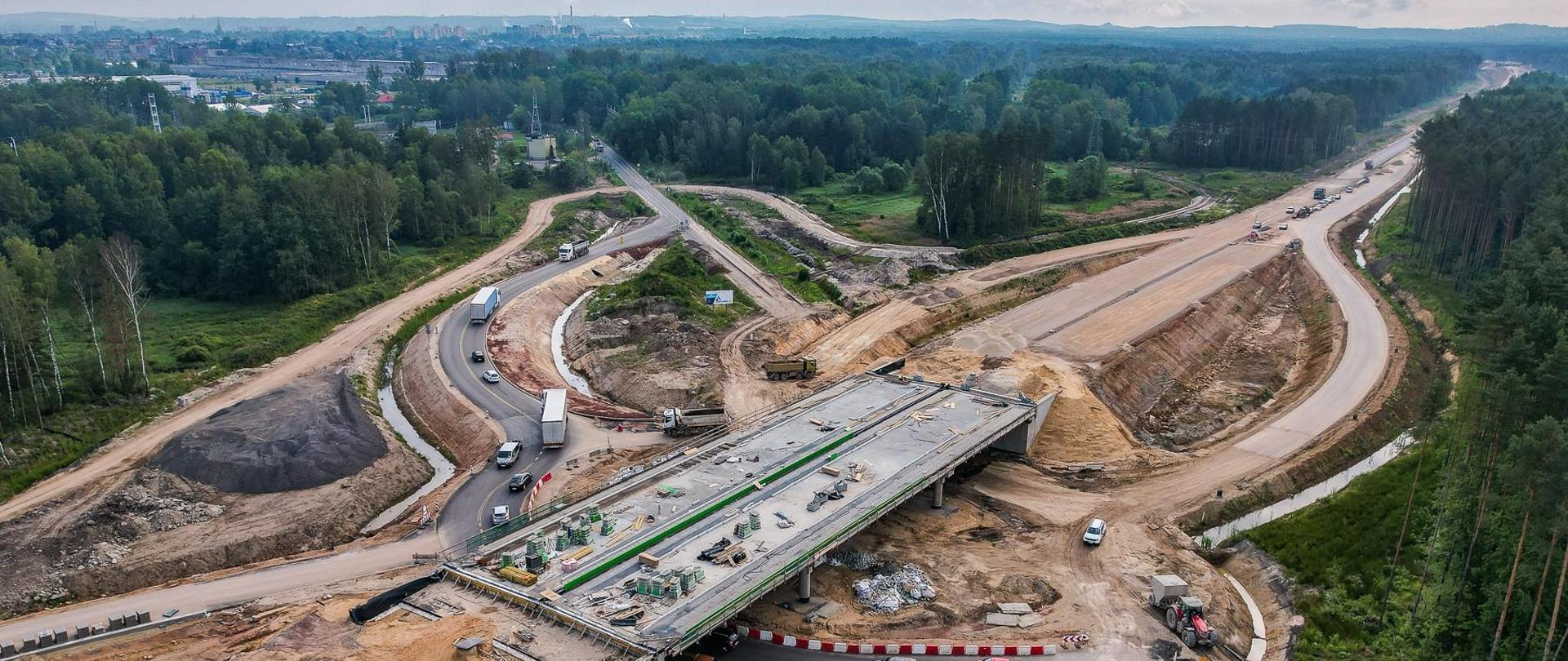 Zdjęcie przedstawia lotniczy widok na duży plac budowy drogi. Widoczny jest częściowo ukończony wiadukt, a pod nim przebiega istniejąca droga. Na miejscu widoczne są maszyny i pojazdy budowlane Otaczający krajobraz obejmuje fragmenty obszarów zalesionych, a w oddali znajduje się zabudowany obszar miasteczka.