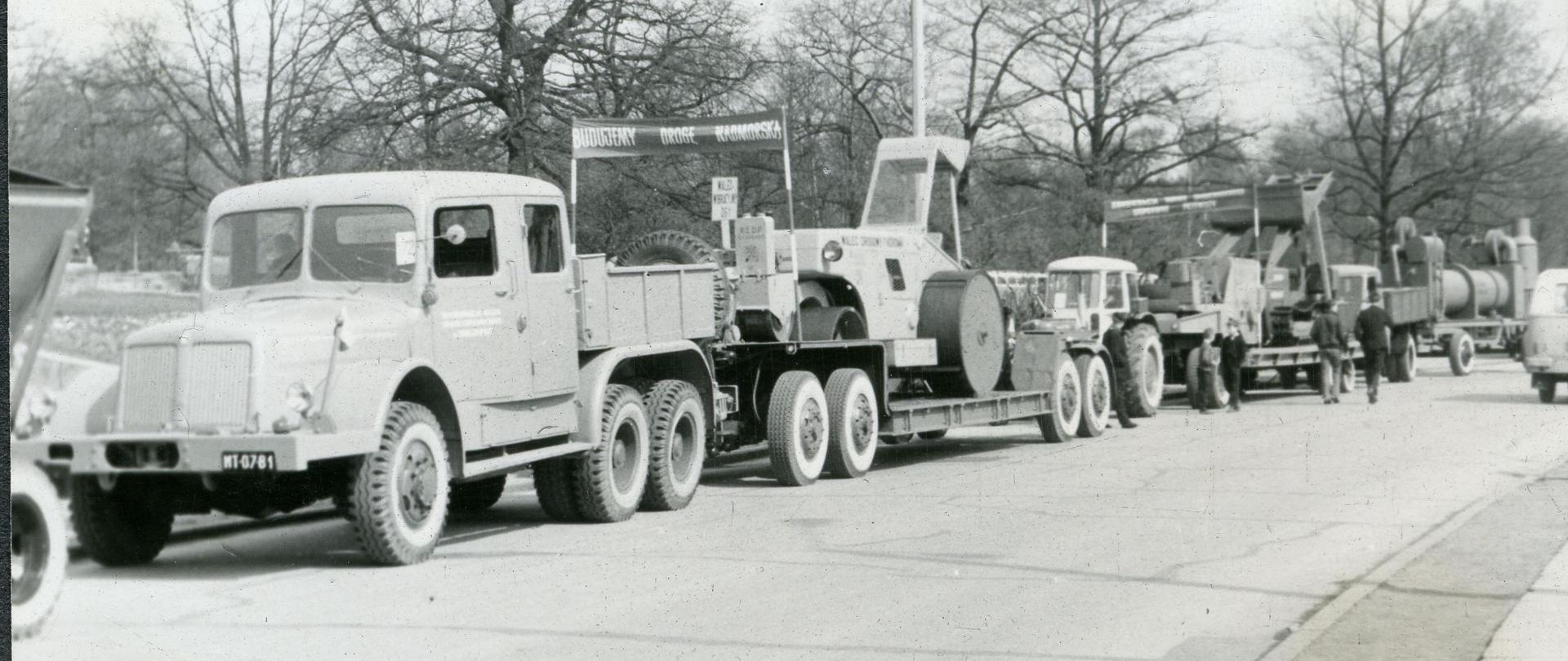 Czarnobiała fotografia. Ustawione na poboczu drogi, przed defiladą, maszyny drogowe. Na czele kolumny samochód ciężarowy z lawetą, na której stoi walec Fadroma WDT 1, dalej w kolumnie ciągnik Ursus, otoczarnia.