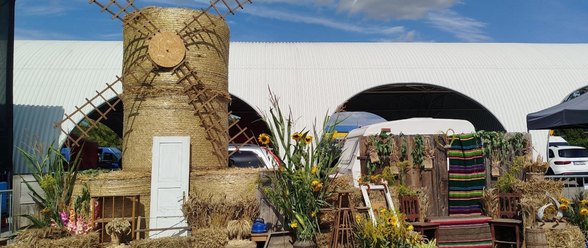 Dekoracja dożynkowa w stylu ludowym – centralnie stoi wiatrak wykonany z bel słomy, otoczony snopami zbóż, słonecznikami i warzywami. Obok ustawiono drewniane krzesła. Dodatkowo położono kolorowe pasiaste nażuty i inne dekoracje. Całość tworzy rustykalny klimat.