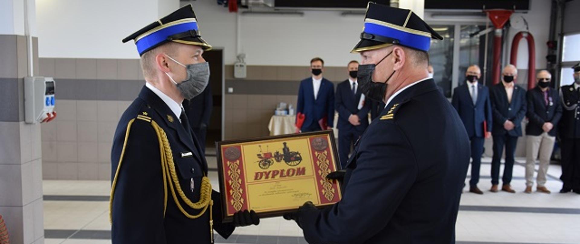 Komendant wojewódzki wręcza dyplom Komendanta Głównego PSP. W tle goście uroczystości.