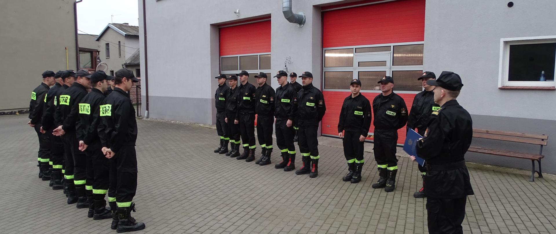 Zdjęcie wykonano w ciągu dnia na placu Komendy Powiatowej Państwowej Straży Pożarnej w Sępólnie Krajeńskim podczas uroczystej zmiany służby obie zmiany służbowe w ubraniach koszarowych stoją w szeregu po lewej i prawej stronie, Na środku na pierwszym planie stoi Komendant powiatowy starszy brygadier Maciej Suchomski w mundurze służbowym.