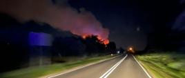 Zdjęcie przedstawia pożar sterty biomasy. Płomienie oraz gęsty dym unoszą się nad stertą biomasy. Zdjęcie zrobione z kabiny samochodu strażackiego, który dojeżdża do miejsca pożaru. 