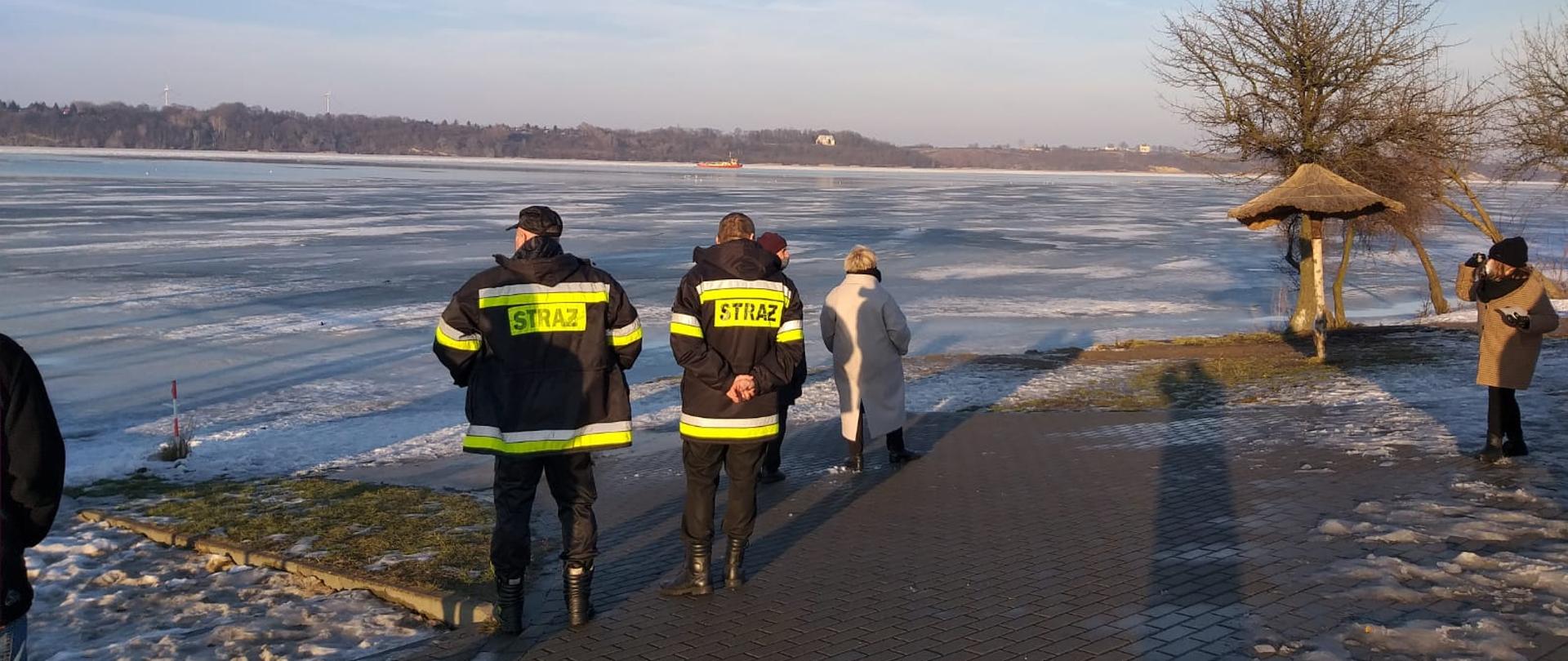 Na zdjęciu dwóch funkcjonariuszy Państwowej Straży Pożarnej zwróconych do rzeki obserwuje na wskaźniku poziom wody. W tle płynie lodołamacz, a w pobliżu przebywa kilka osób ubranych w cywilne stroje.