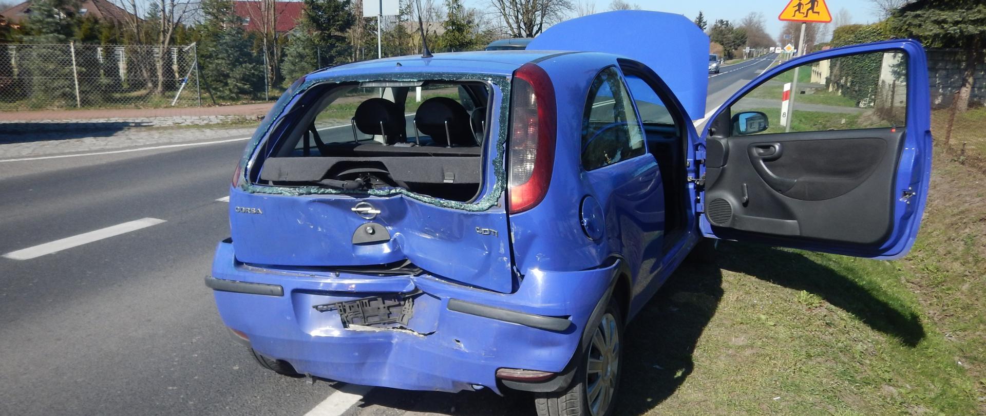 Na zdjęciu widać niebieski samochód Opel Corsa, który uległ uszkodzeniu. Tył pojazdu jest wgnieciony, a tylna szyba wybita. Drzwi po stronie kierowcy są otwarte. Samochód stoi częściowo na poboczu, obok drogi z podwójną linią ciągłą. W tle widać zieloną trawę, drzewa bez liści i domy. Na poboczu stoi również żółty znak drogowy z sylwetkami dzieci. Niebo jest jasne i bezchmurne.