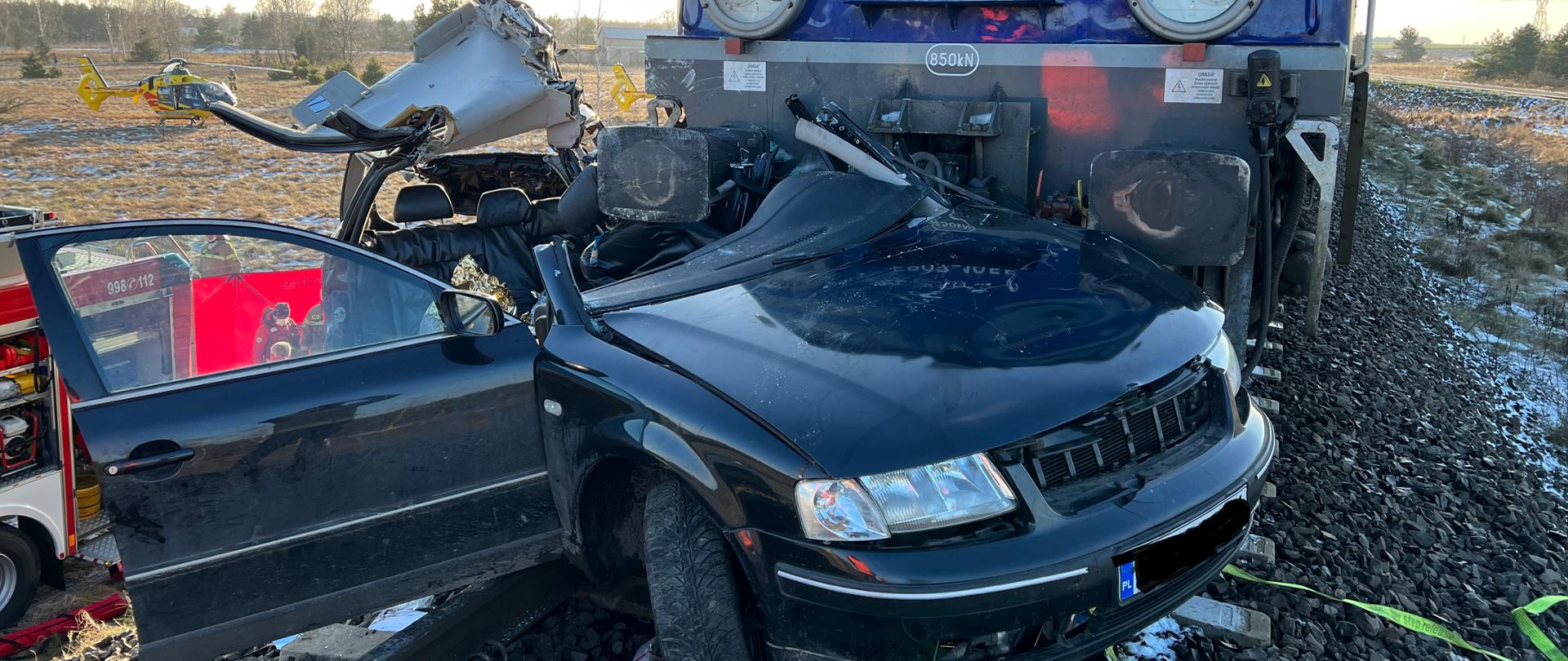 Na torowisku stoi pociąg koloru niebieskiego. Przed nim wrak samochodu osobowego koloru ciemnego po zderzeniu z pociągiem. W oddali widać przyziemiony helikopter ratowniczy LPR. Po lewej stronie przy torowisku fragment wozu strażackiego. Jest pogodnie, świeci słońce.