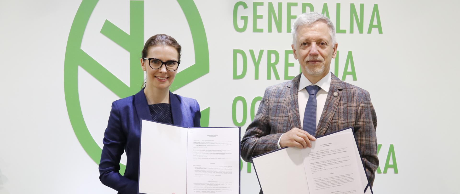 Na tle białej ściany, na której znajduje się logotyp (zielony liść z zielonym napisem Generalna Dyrekcja Ochrony Środowiska) stoi kobieta i mężczyzna. Uśmiechnięta kobieta trzyma w rękach otwartą teczkę. Również mężczyzna, który stoi obok niej trzyma w rękach otwarta teczkę. 