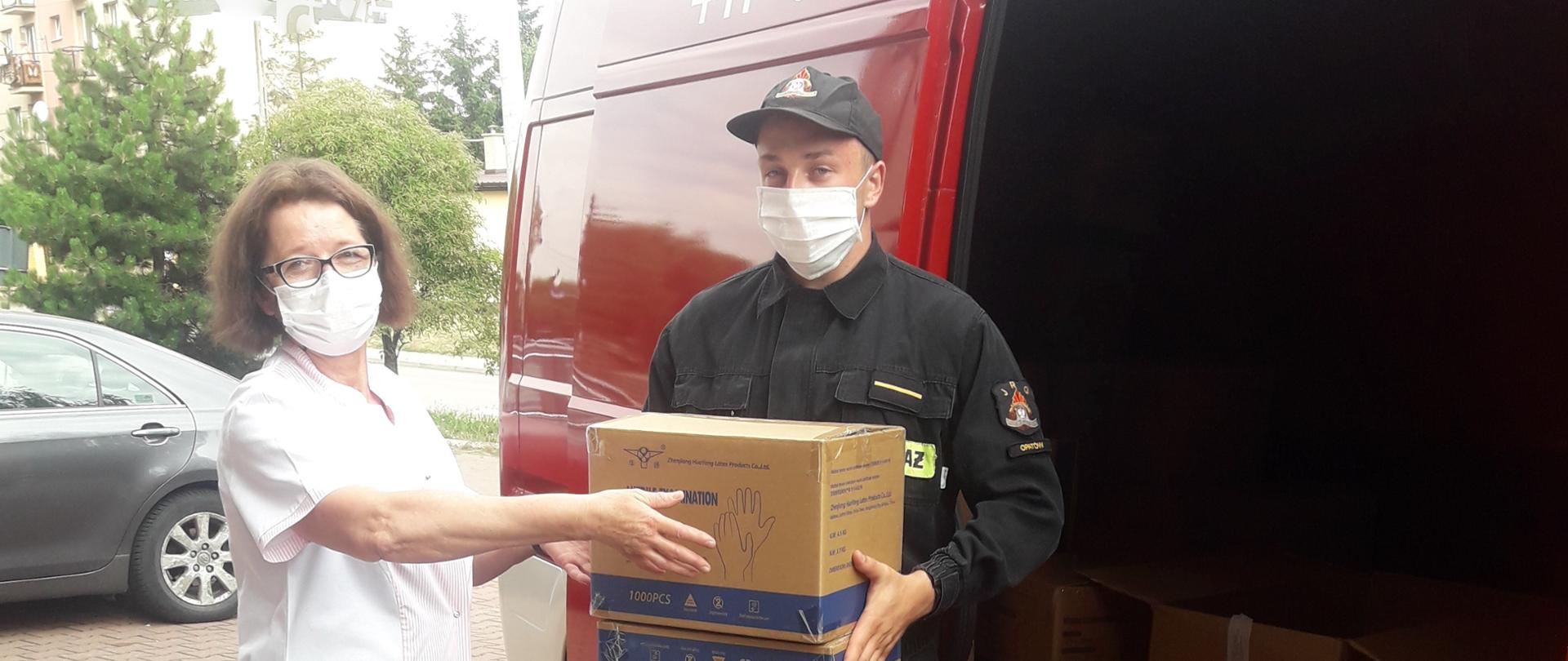 Zdjęcie przedstawia przekazanie Środków Ochrony Indywidualnej przekazywanych służbie ochrony zdrowia przez funkcjonariusza PSP. W tle widać czerwony samochód pożarniczy oraz budynek wielorodzinny.