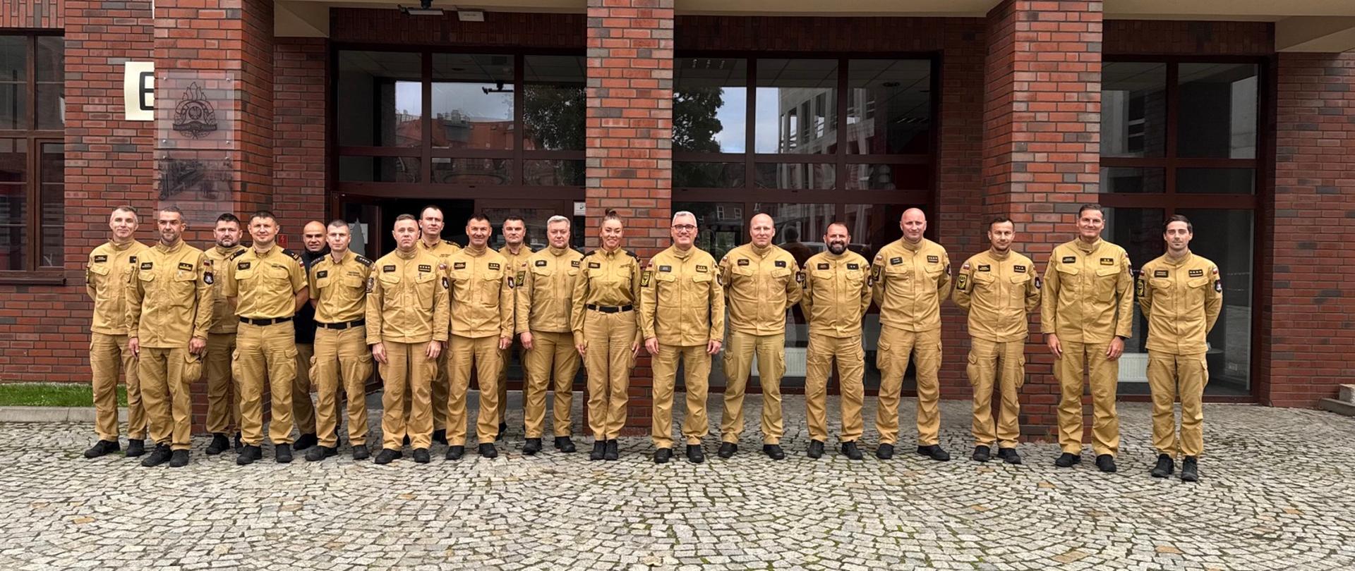 Zdjęcie przedstawia grupę osiemnastu osób ubranych w jasnobrązowe mundury służbowe, stojących w jednym rzędzie na tle ceglanego budynku z dużymi oknami i drzwiami. Przed budynkiem znajduje się plac wyłożony szarą kostką brukową. Wśród stojących osób widać jedną kobietę, która znajduje się mniej więcej w środkowej części grupy. Wszyscy uczestnicy patrzą w stronę aparatu.