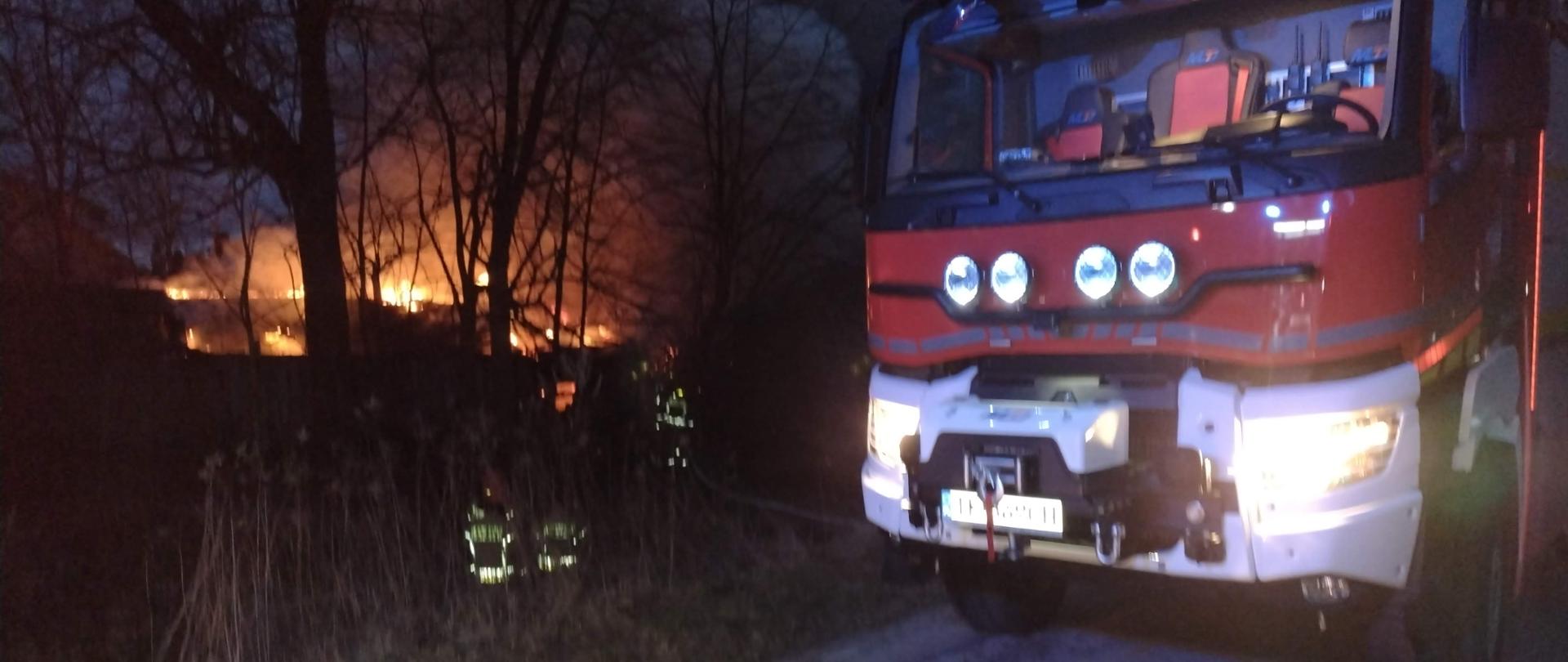 Na zdjęciu widzimy ciężki samochód gaśniczy, palący się budynek gospodarczy, wydobywające się z niego duże ilości dymu oraz strażaków podczas działań gaśniczych.
