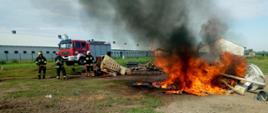 Strażacy stoją trzymając w rękach wąż strażacki zabezpieczając obok palące się palety i wytłaczanki po jajkach. W tle widać samochód strażacki i kurnik.