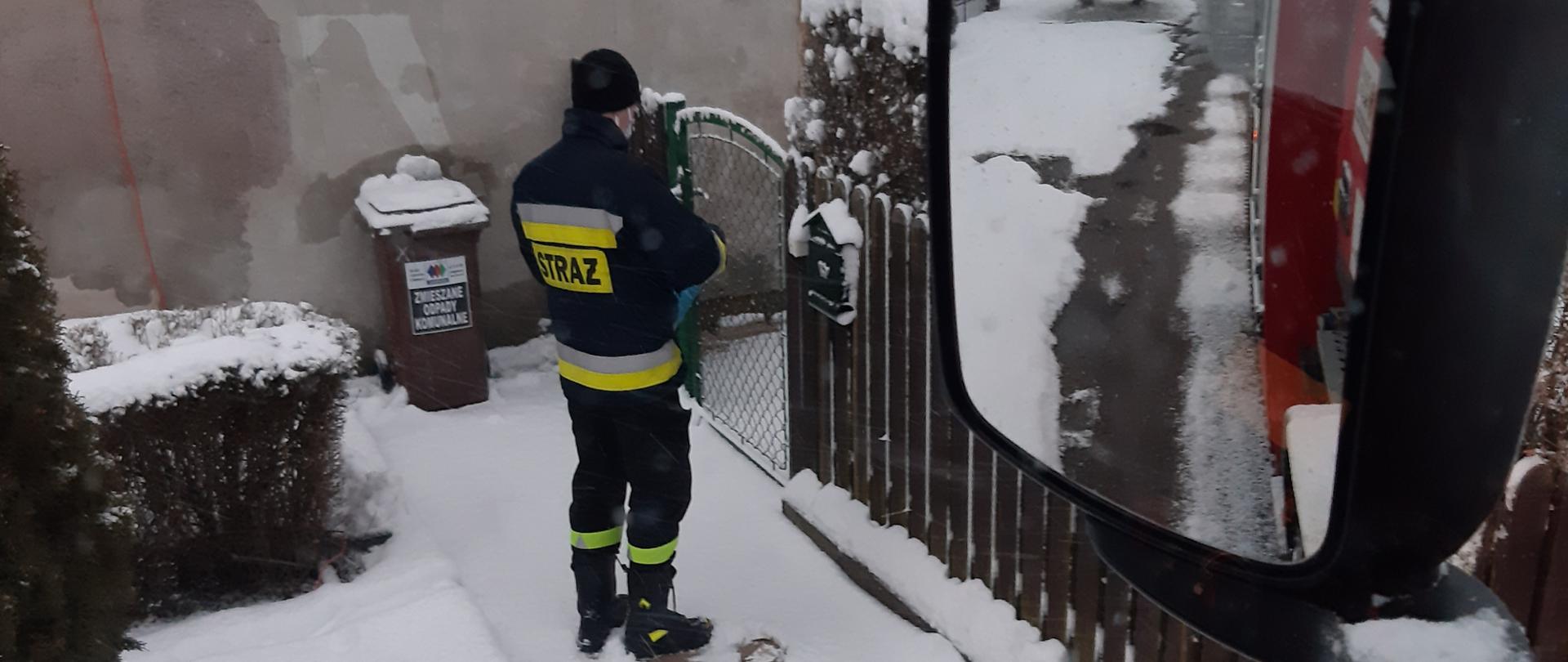 Strażak Ochotniczej Straży pożarnej podczas akcji dostarczania ulotek stoi przed furtką budynku mieszkalnego na ścianie budynku widać liczbę dziesięć. Akcja odbywa się w warunkach zimowych, śnieg znajduje się przed posesją. Mężczyzna odwrócony tyłem do osoby wykonującej zdjęcia. W widocznym na zdjęciu lusterku dostrzegamy bok pojazdu pożarniczego w kolorze czerwonym.