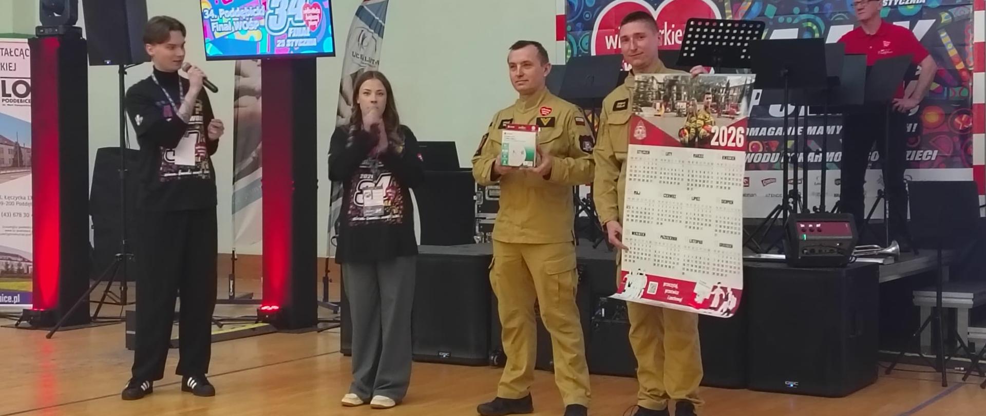 dwaj strażacy stoją na scenie w hali sportowej podczas wydarzenia WOŚP. Jeden trzyma pudełko z urządzeniem (prawdopodobnie czujką dymu), a drugi prezentuje duży kalendarz strażacki na 2026 rok. Towarzyszą im osoby prowadzące z mikrofonami.