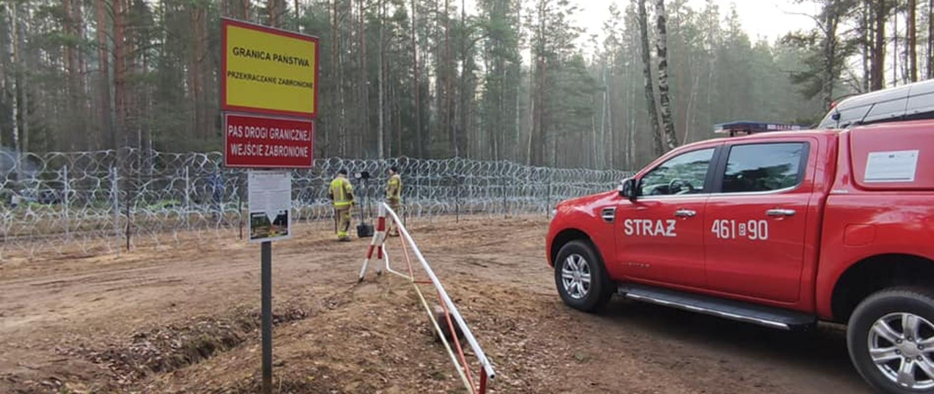 Po prawo widoczny samochód PSP , na wprost widoczna tablica żółto czerwona z napisem GRANICA PAŃSTWOWA, przy drucianej zaporze na granicy dwóch strażaków PSP ustawia oświetlenie
