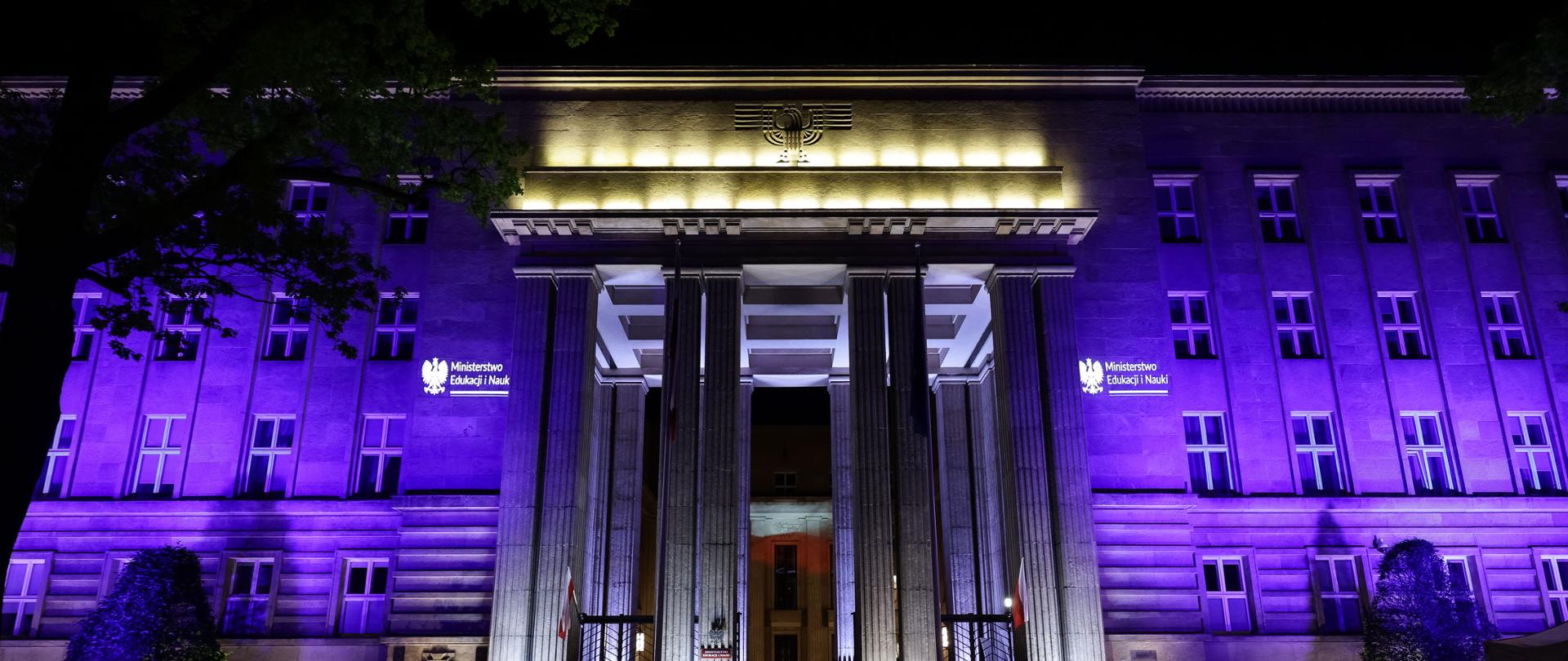 Nocny widok na frontową ścianę budynku ministerstwa, oświetloną na fioletowo po bokach i na żółto nad wejściem.