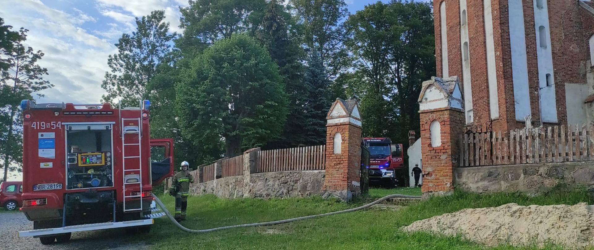 Z prawej strony obiekt kościoła z cegły, ogrodzenie z kamieni i desek. Przez bramę wejściową widoczne auto strażackie. Z lewej strony samochód strażacki i strażak. W oddali zielone drzewa.