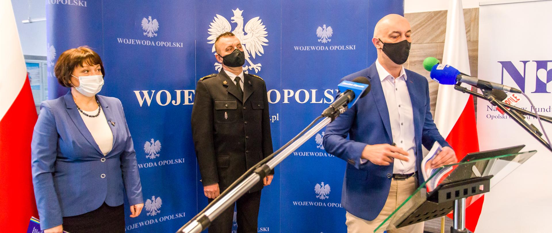 Zdjęcie konferencji prasowej Wojewody Opolskiego. Od lewej strony stoi Pani Dyrektor Wojewódzkiej Stacji Sanitarno Epidemiologicznej w niebieskim żakiecie i czarnej spódnicy z białą maseczką na twarzy. Następnie 1,5 metra po jej lewej stronie stoi na tle ścianki z napisem "Wojewoda Opolski" i białym godłem Polski, w ubraniu wyjściowym st. bryg. Arkadiusz Kuśmierski Zastępca Opolskiego Komendanta Wojewódzkiego PSP z czarną maseczką na twarzy. Przed Komendantem przy mównicy stoi Adrian Czubak Wojewoda Opolski z niebieskiej marynarce, czarnej maseczce na twarzy i jasnych spodniach. Przed Wojewodą widoczne trzy mikrofony. W tle dwie flagi Polski oraz po prawej stronie kawałek roll up z napisem "NFZ".