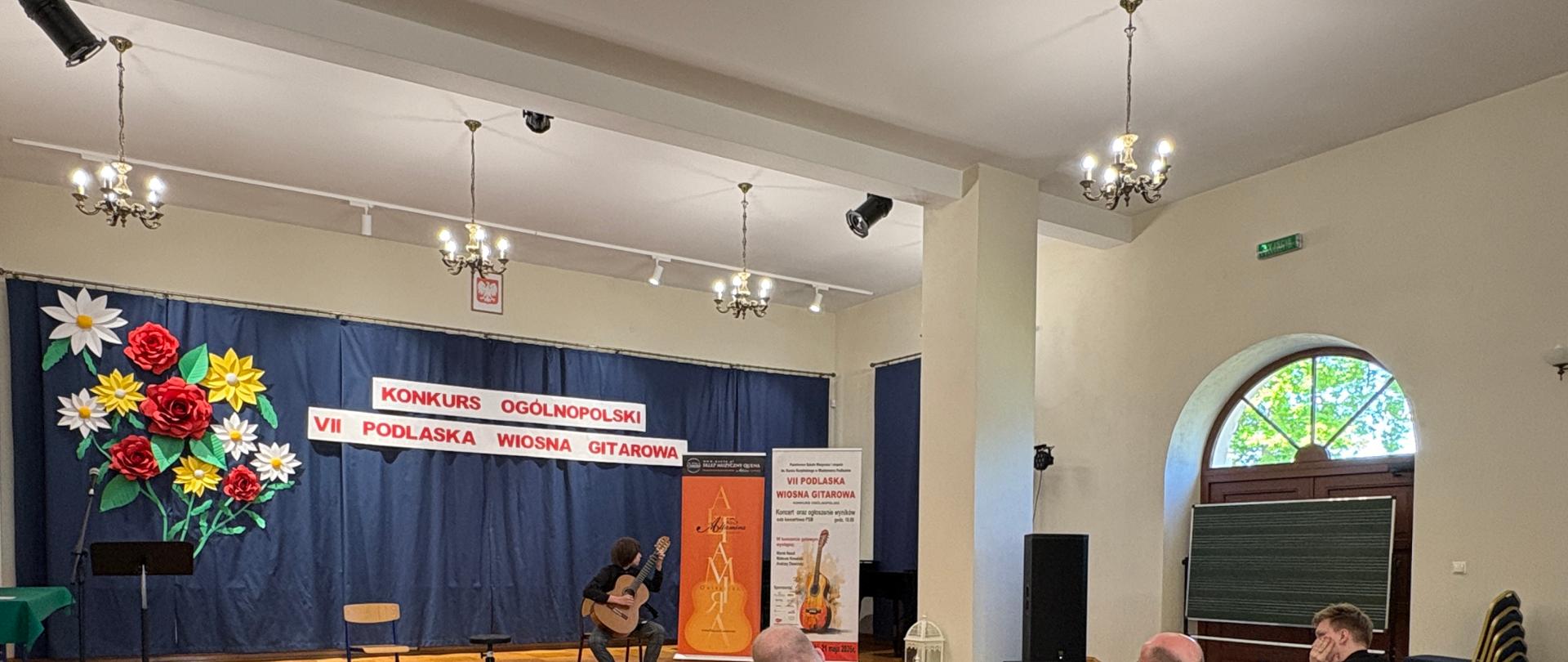 21 maja 2026 roku w Międzyrzecu Podlaskim odbył się ogólnopolski konkurs gitarowy „VII Podlaska Wiosna Gitarowa”. Naszą szkołę reprezentowali uczniowie klasy gitary pani Iwony Grabowskiej – Jan Szabat z klasy III cyklu sześcioletniego wywalczył II miejsce w kategorii I.
Jan Szabat na scenie podczas występu konkursowego. Ubrany elegancko, siedzi w pozycji koncertowej, skupiony na wykonaniu swojego programu artystycznego. Obok niego znajduje się podnóżek gitarowy oraz stołek. W tle widoczne są banery konkursowe z napisem „Konkurs Ogólnopolski” oraz „VII Podlaska Wiosna Gitarowa”. Po prawej stronie sceny znajduje się również reklama gitar klasycznych Altamira. Zdjęcie oddaje atmosferę konkursu muzycznego. Widać jurorów siedzących przed sceną.
