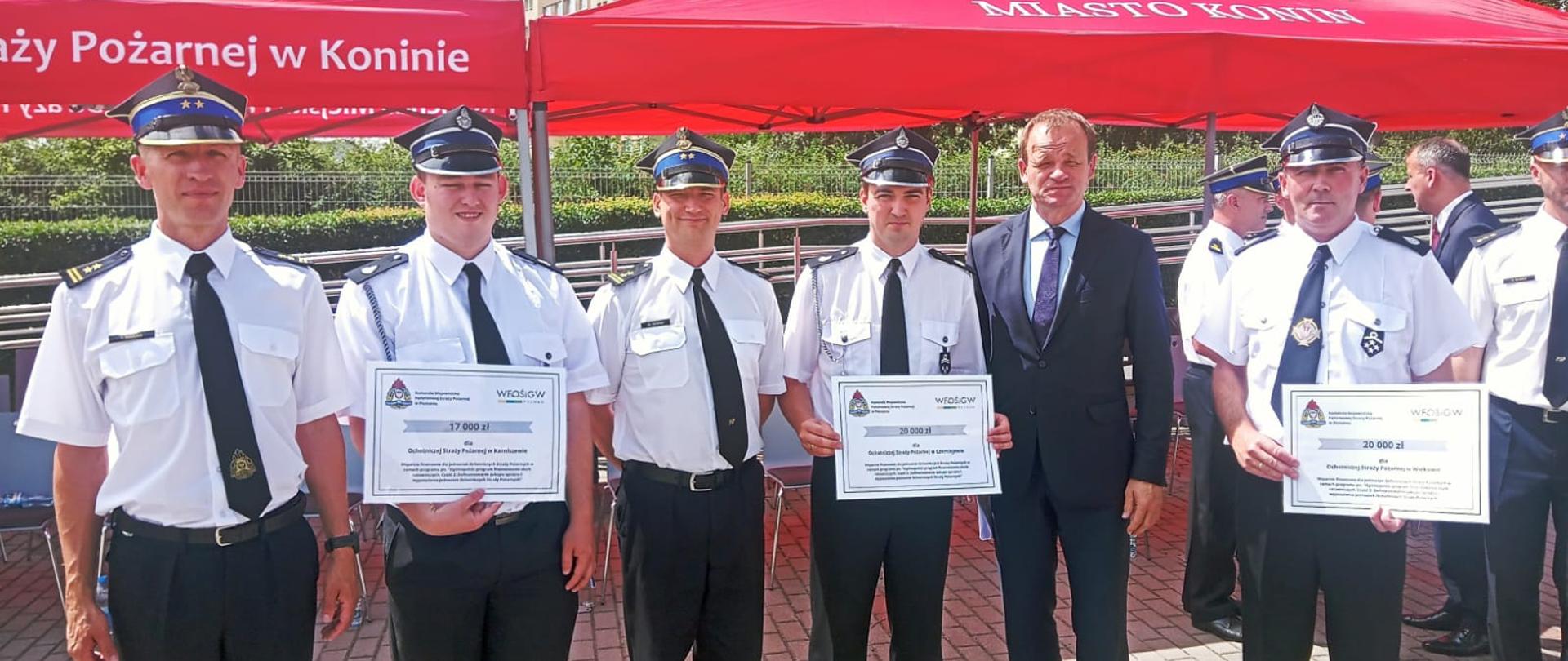 Grupa strażaków w mundurach galowych - czarne spodnie, biała koszula z czarnym krawatem i strażacka rogatywka stoi wraz z osobą w czarnym garniturze. Trzech strażaków trzyma w rękach karty z promesą.W tle czerwone namioty bez ścian oraz wysoki budynek mieszkalny i zielone drzewa