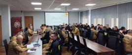 Na zdjęciu w sali przy stolach siedzą strażacy w mundurach, na ścianie wyświetlona prezentacja multimedialna