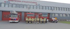 Strażacy z KP PSP Opatów w szeregu przed samochodami bojowymi ustawionymi na placu zewnętrznym budynku Komendy. Samochody z włączonymi sygnałami świetlnymi.