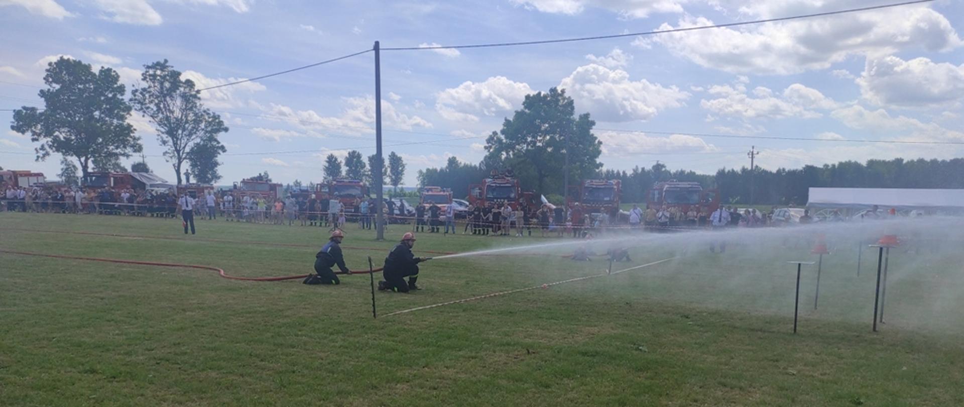 Zawodnicy strumieniem wody strącają stożki ze stojaków. 