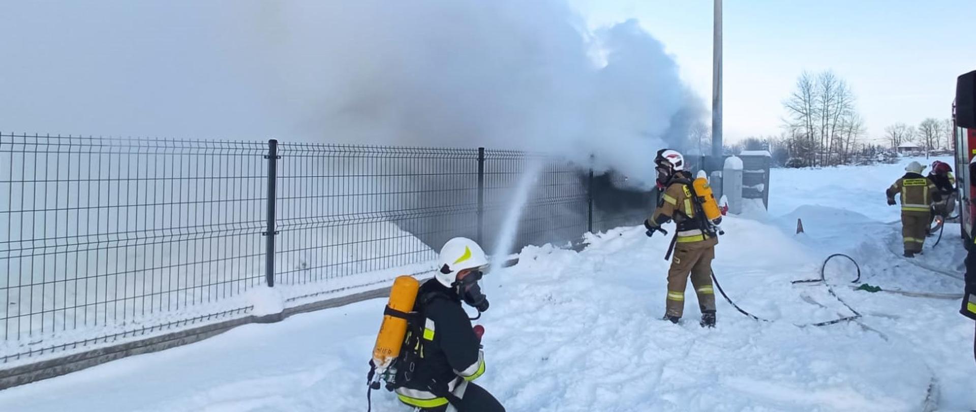 Na zdjęciu: panelowe ogrodzenie, za którym w centralnym punkcie, w kłębach dymu i pary wodnej stoi budynek biurowy. Przed ogrodzeniem dwóch strażaków w aparatach ochrony dróg oddechowych: jeden w pozycji kucznej podaje wodę na palący się obiekt , drugi z wężem szybkiego natarcia przygotowuje się do gaszenia. Po prawej stronie widać jeszcze dwóch ratowników.