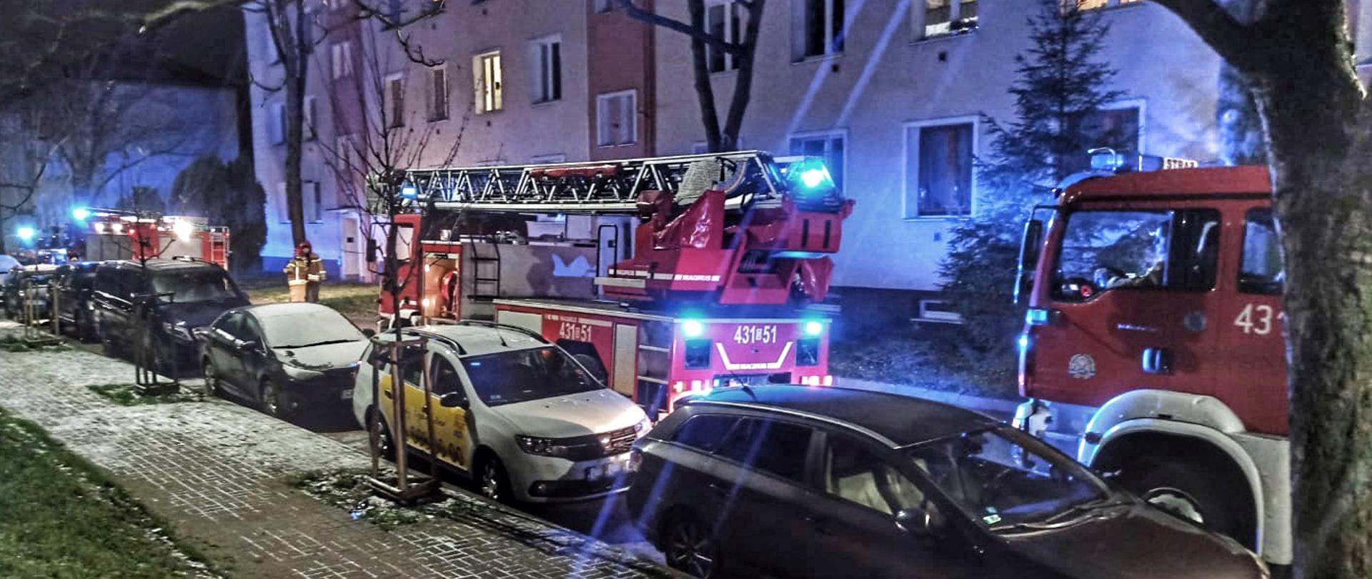 Pojazdy pożarnicze stoją na wąskiej ulicy wzdłuż zaparkowanych pojazdów osobowych przed trzykondygnacyjnym budynkiem.