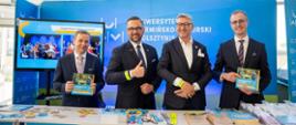 Przedstawiciele uczelni i instytucji edukacyjnych pozują do zdjęcia przy stoisku promocyjnym Uniwersytetu Warmińsko-Mazurskiego w Olsztynie podczas targów edukacyjnych. Na pierwszym planie widoczne są materiały informacyjne dla kandydatów na studia, gadżety promocyjne oraz informatory rekrutacyjne. W tle znajduje się ekspozycja uczelni z ekranem prezentującym działalność akademicką i ofertę edukacyjną.