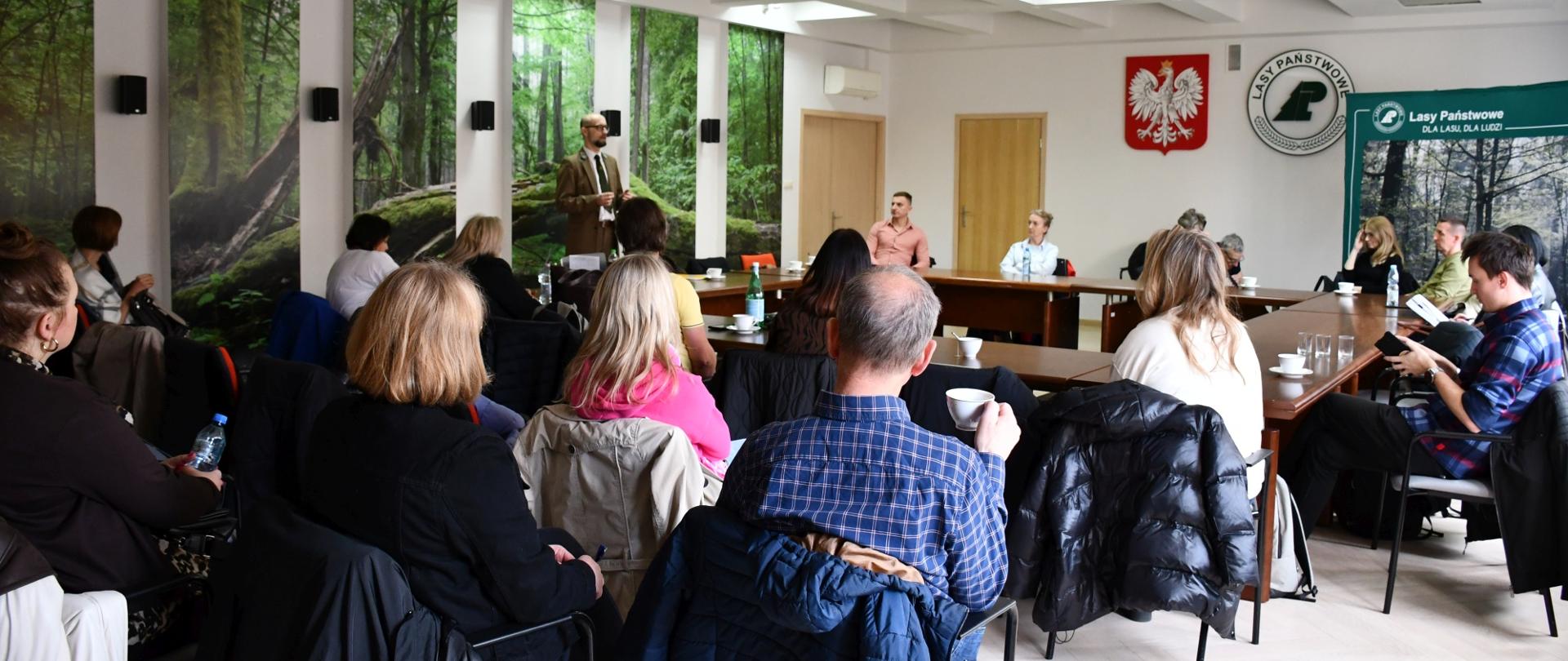 Sala, na ścianach zdjęcia lasu, godło i logo Lasów Państwowych, widok z tyłu na ludzi siedzących na krzesełkach, za stołem komisja konkursowa