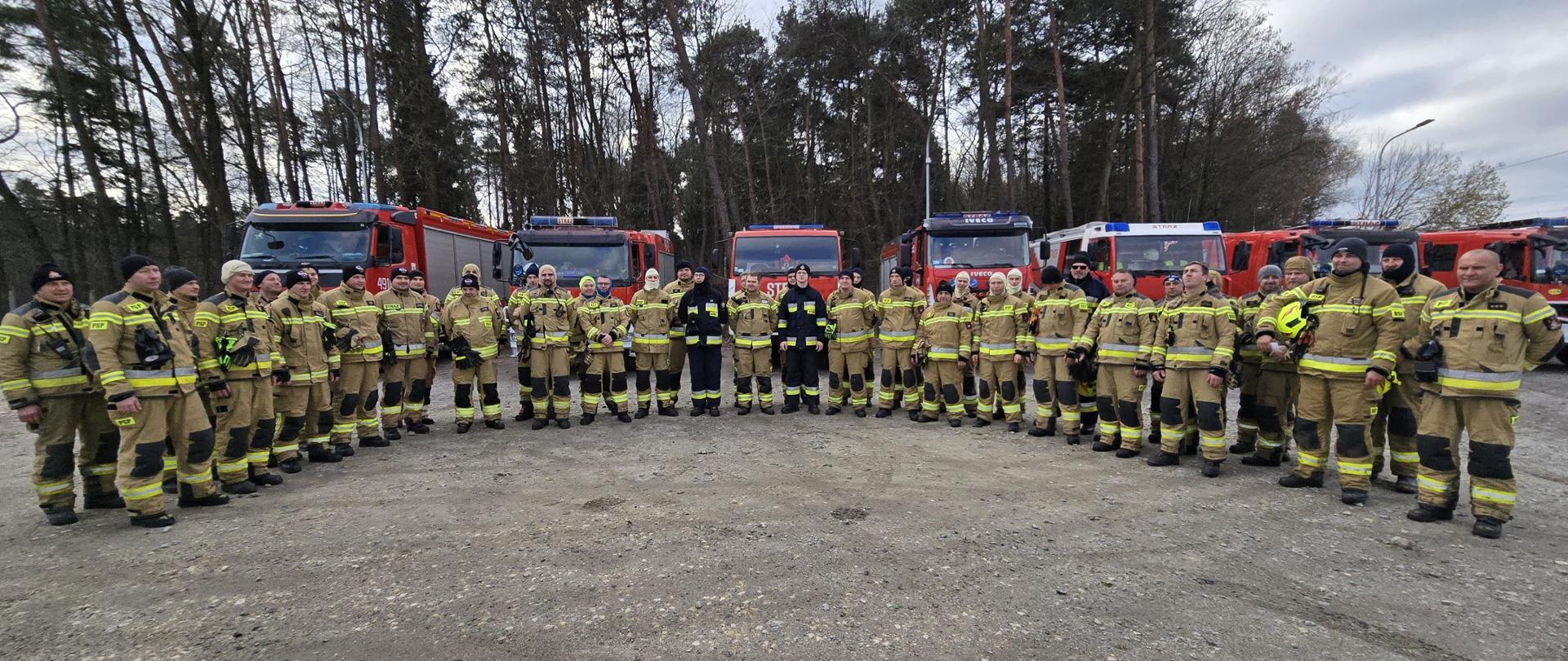 Na zdjęciu znajdują się strażacy w ubraniach specjalnych ustawieni przed pojazdami strażackimi