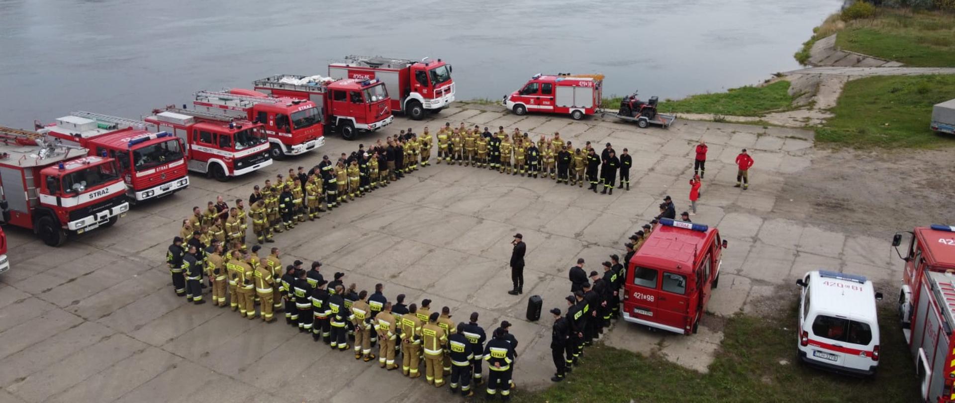 Na zdjeciu widać omówienie ćwiczeń strażackich przez Komendanta. Ochotnicy stoją w kwadracie w dwu szeregu a obok nich znajduja sie wozy strazackie.