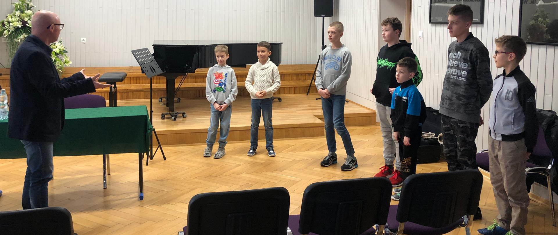 Po lewej stronie Pedagog mówiący do grupy uczniów stojących na przed nim po prawej stronie. W tle – czarny fortepian oraz pulpit, logo szkoły oraz Godło.