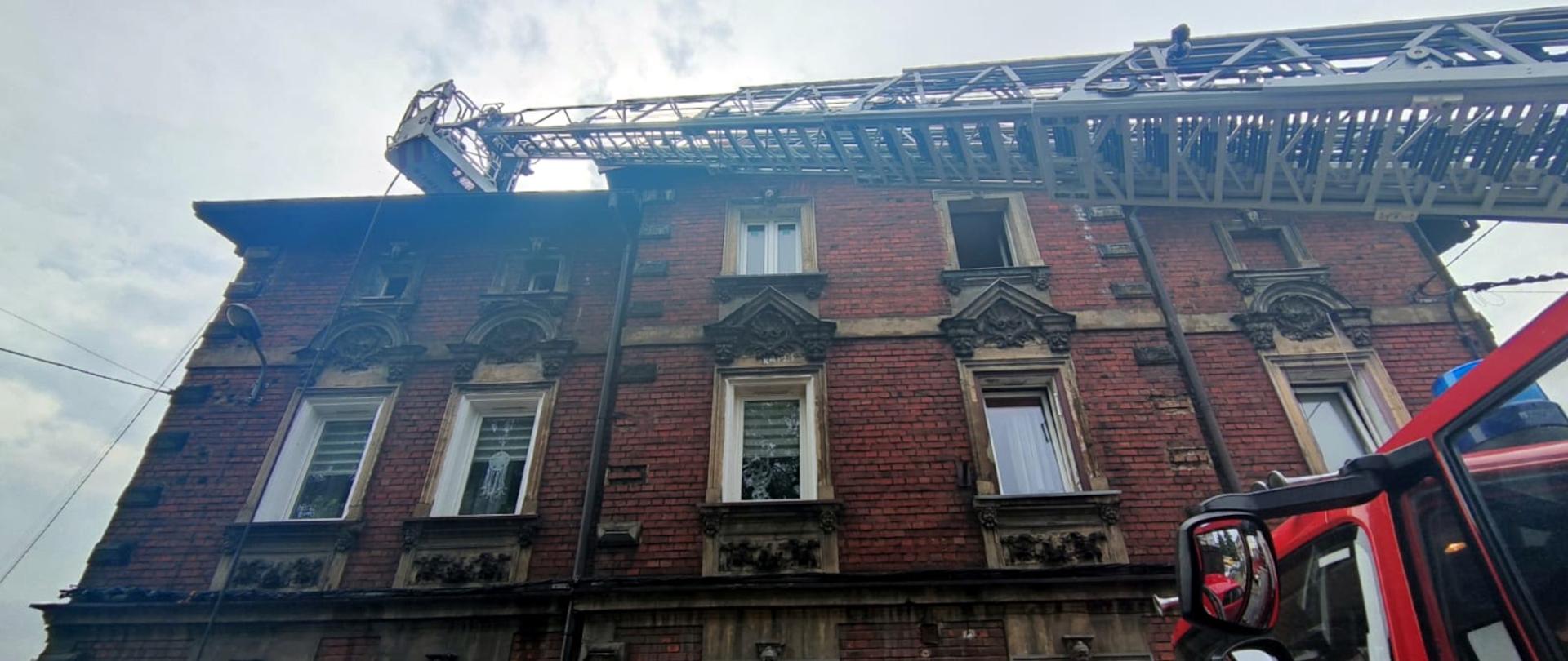 Zdjęcie przedstawia elewację palącego się budynku. Do dachu przystawiona jest drabina mechaniczna. 