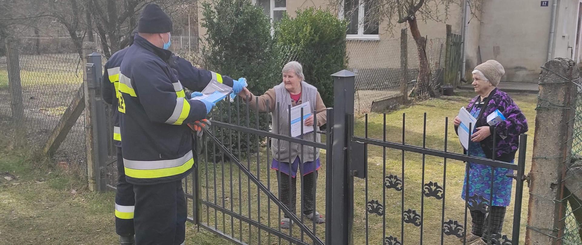 Zdjęcie przedstawia dwóch strażaków z OSP Nowa Niedrzwica przekazujących ulotki oraz maseczki ochronne mieszkankom Nowej Niedrzwicy