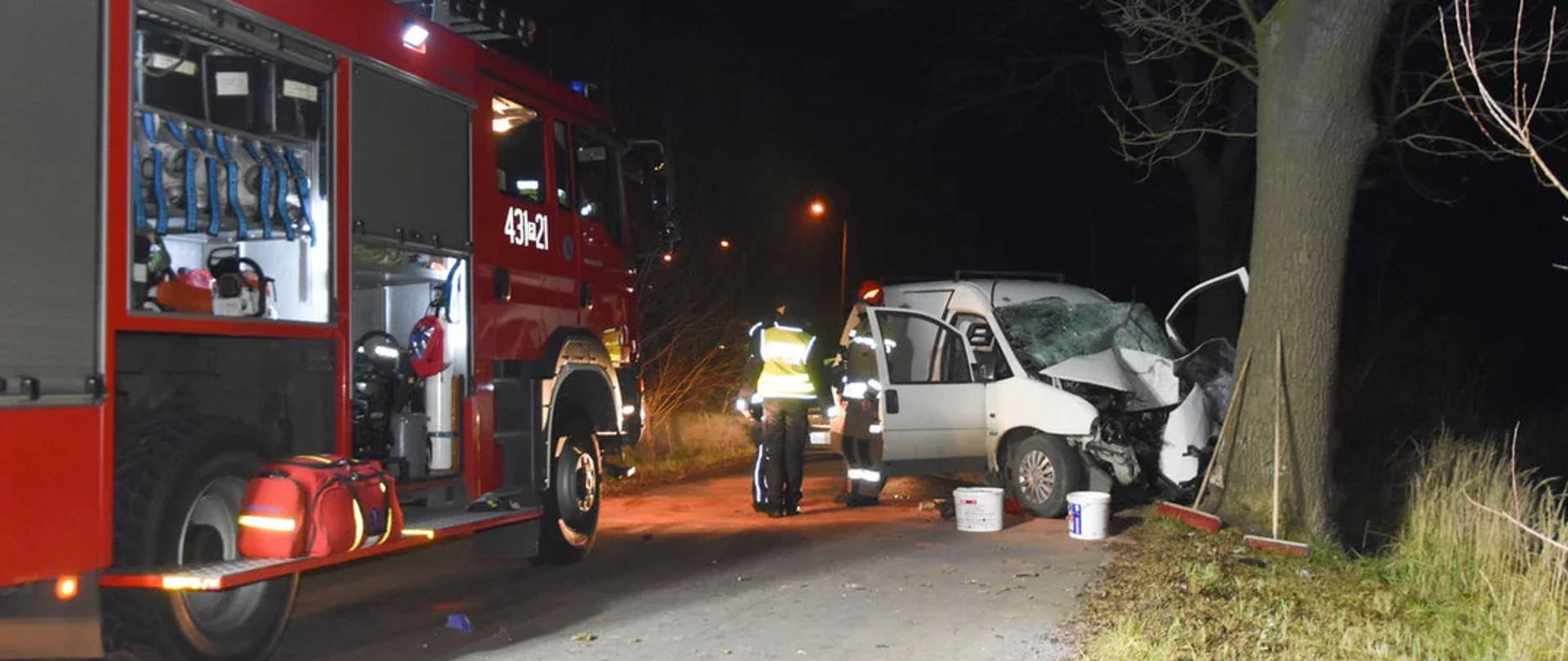 Samochód osobowy stoi na drodze, po uderzeniu w drzewo. W tle samochód pożarniczy.