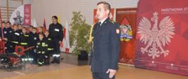 Wręczenie promes. Sala sportowa w Szkaradowie. Na te materiałów promujących KP PSP w Rawiczu stoi strażak PSP w umundurowaniu wyjściowym, pełniący rolę dowódcy uroczystości. Z lewej strony członkowie MDP w Ostojach przy sikawce konnej