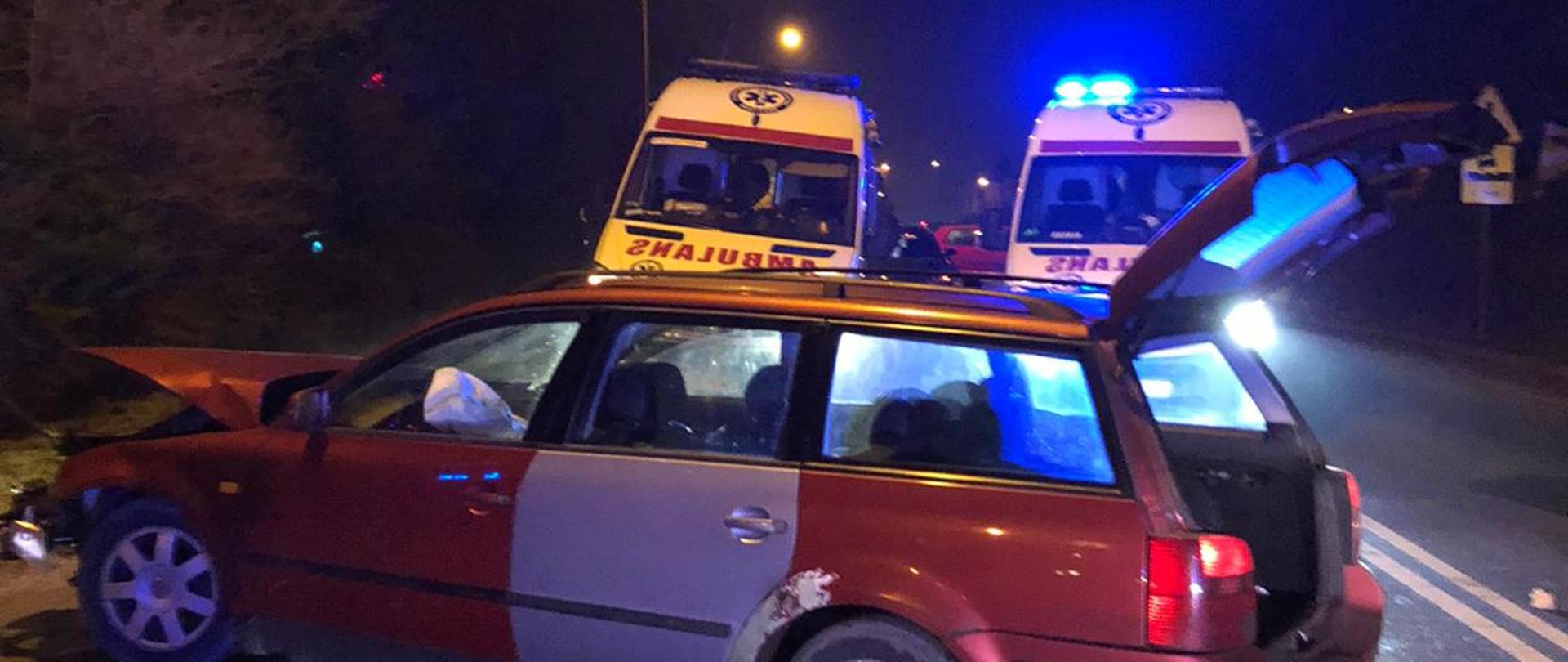 Zdjęcie przedstawia czerwony pojazd marki Volkswagen z rozbitym przodem i otwartym bagażnikiem, który stoi na drodze asfaltowej oraz chodniku dla pieszych. Na drugim planie znajdują się dwa zespoły ratownictwa medycznego. Zdjęcie wykonane po zmierzchu słońca.