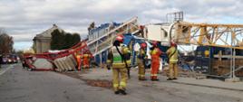 Na zdjęciu widoczny przewrócony żuraw wieżowy na kontenery pracownicze. Przed nim strażacy.