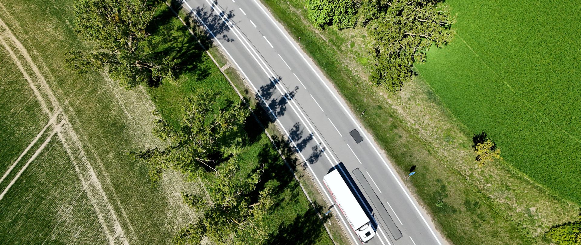 Zdjęcie przedstawia widok z lotu ptaka na biały ciężarówkę jadącą po dwupasmowej drodze otoczonej zielonymi polami i drzewami. Droga ma wyraźne oznaczenia pasów, a cienie drzew padają na jezdnię. Sceneria znajduje się na obszarze wiejskim z dobrze utrzymaną roślinnością