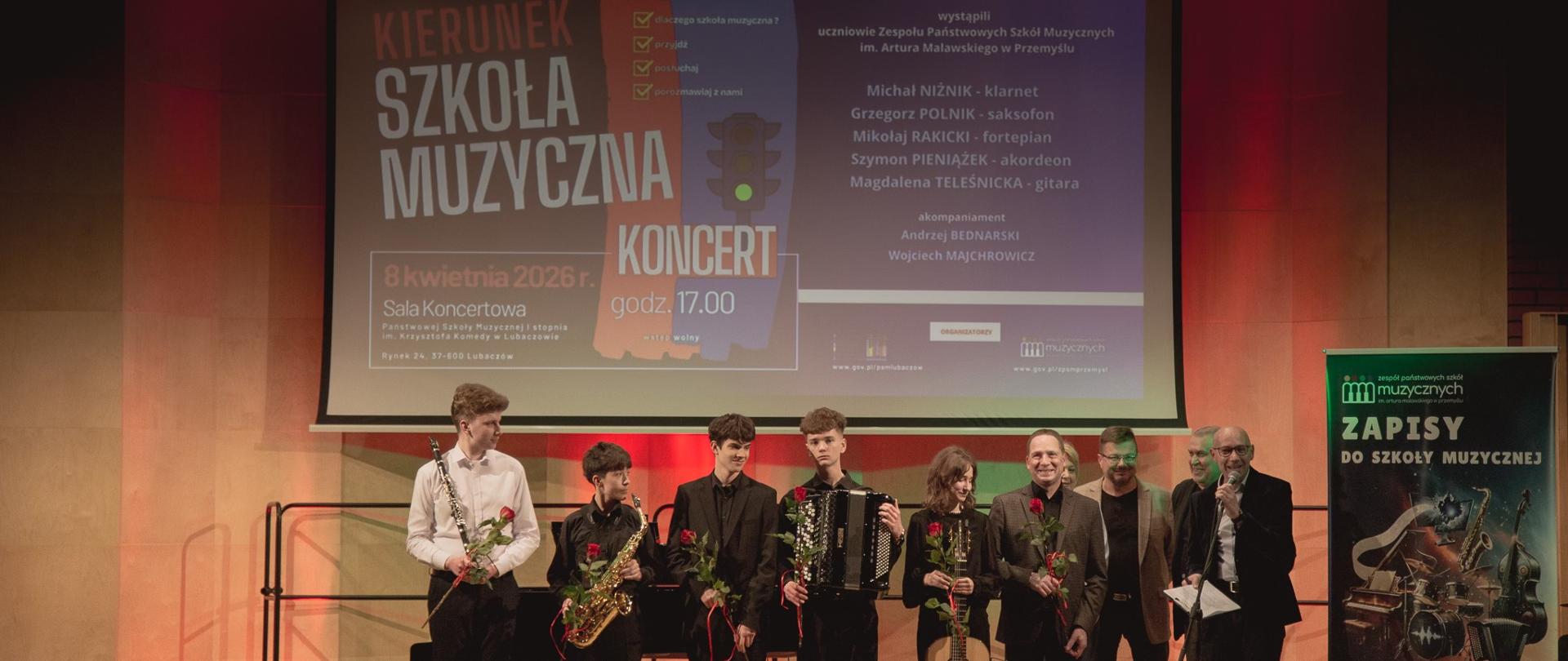 Zdjęcie dokumentuje finał koncertu zatytułowanego „Kierunek Szkoła Muzyczna”, który odbył się 8 kwietnia 2026 roku w Sali Koncertowej Państwowej Szkoły Muzycznej im. Krzysztofa Komedy w Lubaczowie. Na pierwszym planie, w ciepłym blasku czerwonego oświetlenia scenicznego, stoi grupa pięciu młodych muzyków – uczniów Zespołu Państwowych Szkół Muzycznych im. Artura Malawskiego w Przemyślu. Występujący, ubrani w eleganckie, ciemne stroje, prezentują swoje instrumenty: klarnet, saksofon, akordeon oraz gitarę klasyczną, a każdy z nich trzyma w dłoni symboliczną czerwoną różę.
W tle, nad głowami artystów, widnieje duży ekran z plakatem informacyjnym, który wymienia nazwiska wykonawców: Michała Niżnika, Grzegorza Polnika, Mikołaja Rakickiego, Szymona Pieniążka oraz Magdaleny Teleśnickiej, a także akompaniatorów – Andrzeja Bednarskiego i Wojciecha Majchrowicza. Po prawej stronie sceny towarzyszy im grupa dorosłych mężczyzn, prawdopodobnie organizatorów lub pedagogów, z których jeden przemawia do mikrofonu, domykając część oficjalną wydarzenia. Całość kompozycji dopełnia baner zachęcający do zapisów do szkoły muzycznej, co podkreśla promocyjny i edukacyjny charakter tego artystycznego spotkania.