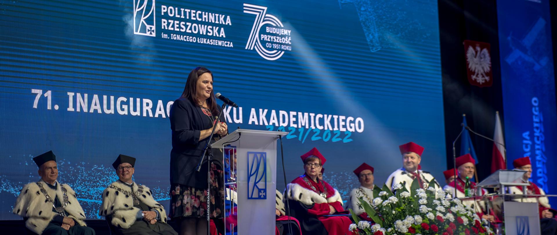 wiceminister Małgorzata Jarosińska-Jedynak mówi do mikrofonu, za nią siedzą profesorowie Politechniki Rzeszowskiej, napis na ścianie: 71 inauguracja roku akademickiego Politechnika Rzeszowska im. Ignacego Łukasiewicza, 70 lat budujemy przyszłość