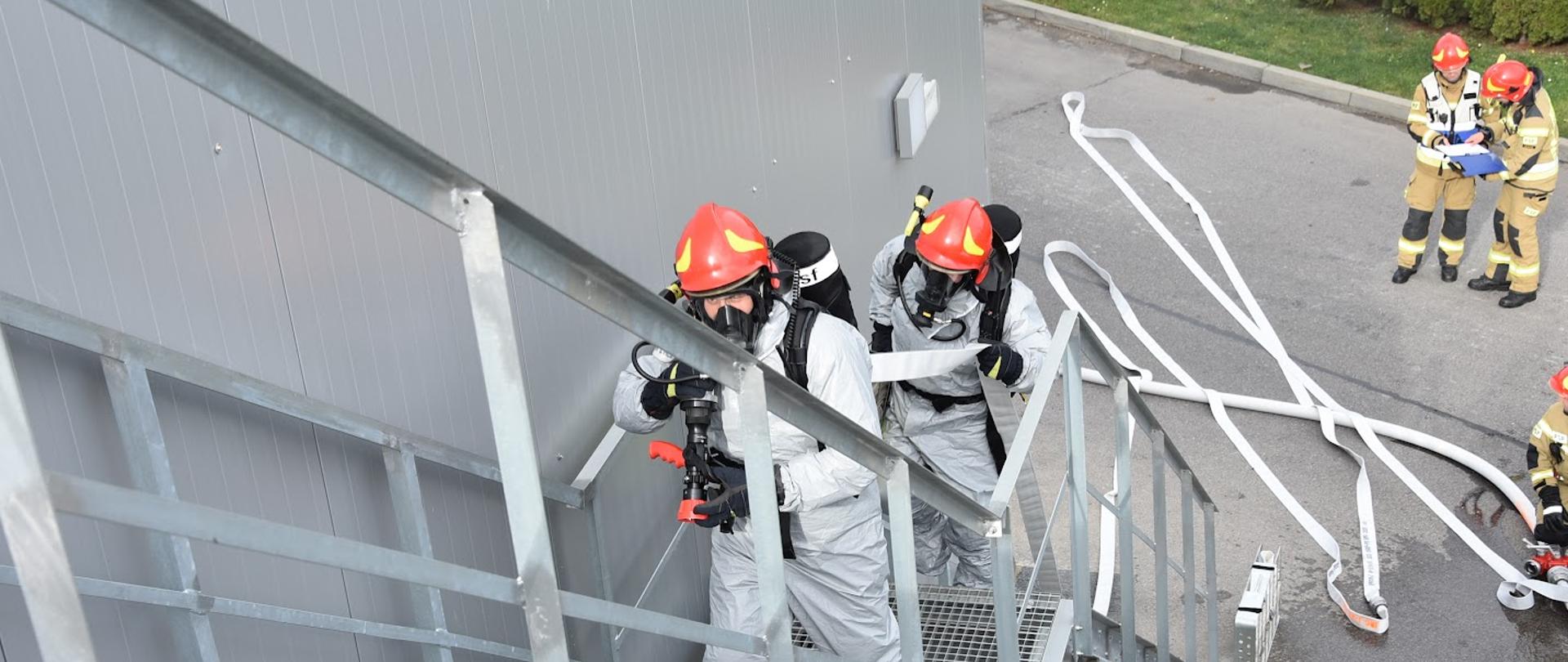 Zdjęcie przedstawia dwóch strażaków ubranych w ubrania przeciwochlapaniowe i sprzęt ochrony układu oddechowego, którzy wchodzą po zewnętrznej klatce schodowej z linią gaśniczą. W tle pracują inni strażacy.