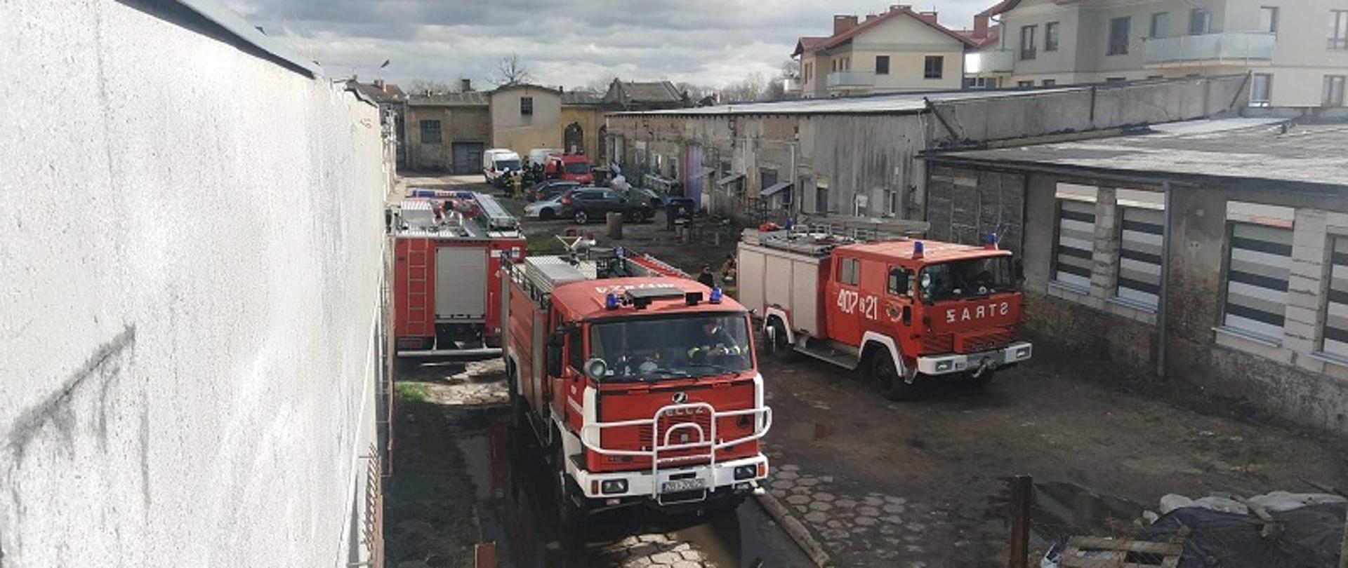 Zdjęcie przedstawia plac wewnętrzy zakładu w którym doszło do pożaru poddasza w parterowym budynku magazynowo-warsztatowym. Na pierwszym planie trzy czerwone samochody pożarnicze. W głębi po prawej stronie obiekt objęty działaniami gaśniczymi z dwójką strażaków znajdujących się na dachu w pozycji klęczącej